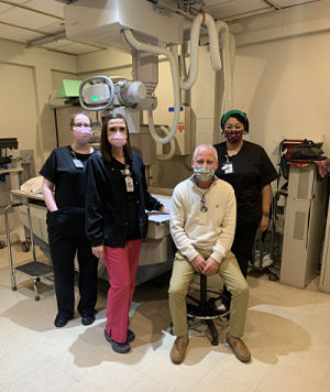 Picture of a Physician (Male) with Nurses (Females) wearing mask in an Imaging Room. There is a bed with a CT scan in the room